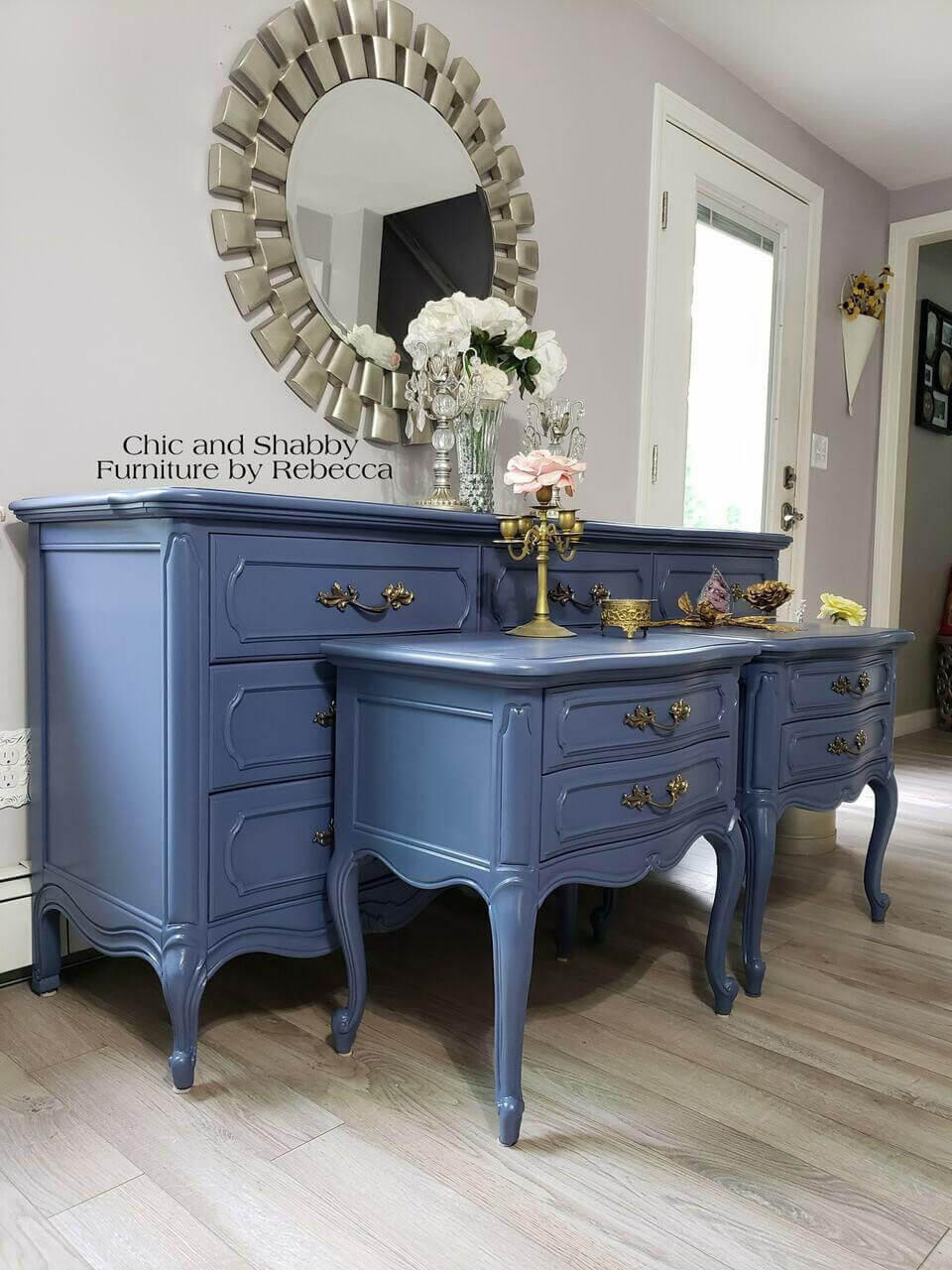 Dixie Belle Paint Yankee Blue Chalk Mineral Paint is the perfect paint for any DIY project!  A Yankee Blue chalk painted dresser and two matching blue chalk painted night stands sit on a light brown floor, in front of a gray wall with white trim.  A round mirror in a silver frame hang on the wall above the blue dresser.  A white and pink flowers, a clear vase, a bronze candlestick and other decorations sit on top of the chalk painted pieces.