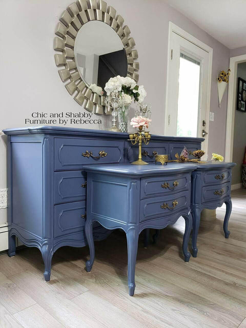 Dixie Belle Paint Yankee Blue Chalk Mineral Paint is the perfect paint for any DIY project!  A Yankee Blue chalk painted dresser and two matching blue chalk painted night stands sit on a light brown floor, in front of a gray wall with white trim.  A round mirror in a silver frame hang on the wall above the blue dresser.  A white and pink flowers, a clear vase, a bronze candlestick and other decorations sit on top of the chalk painted pieces.