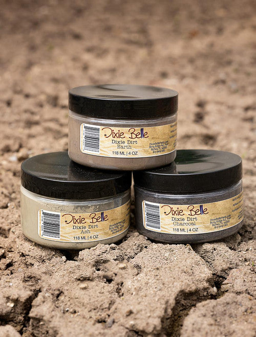 Three jars of Dixie Belle Dixie Dirt in different shades are stacked on dirt, with black lids and tan labels, each containing 118 ml (4 oz) of product. The background is a textured soil surface.