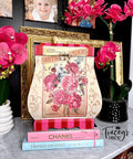 A decorative setup featuring a lamp-shaped floral art piece with "Vintage Flowers" text, framed photos, and pink orchids. Below are two books: "Chanel," and "Things We Love."