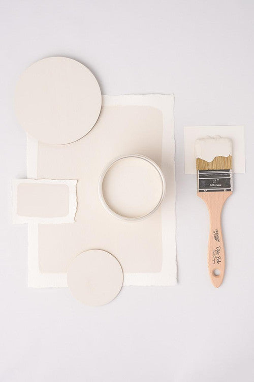 White paint samples arranged artfully with a jar of paint and a wooden-handled paintbrush on a white background. The samples include circular and rectangular pieces of paper.