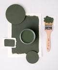 A flat lay image featuring a jar of green paint on textured paper, surrounded by painted circles, a paintbrush with green paint on the bristles, and swatches. The composition is arranged neatly on a white background.