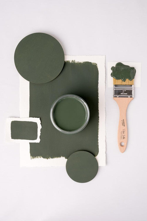 A flat lay image featuring a jar of green paint on textured paper, surrounded by painted circles, a paintbrush with green paint on the bristles, and swatches. The composition is arranged neatly on a white background.