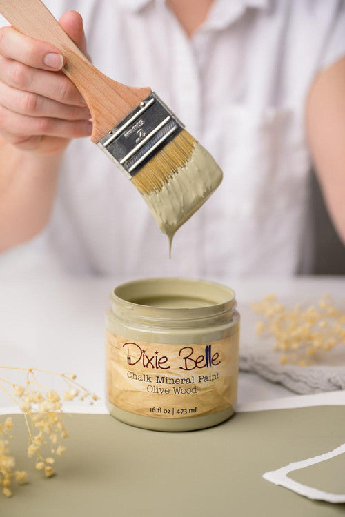 Person holding a brush with greenish paint dripping over an open jar labeled "Dixie Belle Chalk Mineral Paint Olive Wood," placed on a surface with dried flowers nearby.