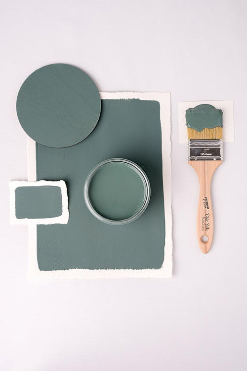 A flat lay image featuring a jar of green paint, a paintbrush with a wooden handle partially covered in green paint, and green paint swatches on white paper, all against a light background.