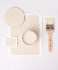 A flat lay image shows a beige paint theme with a round board, a paint can, and two swatches on white paper. A wooden-handled paintbrush with beige paint is placed to the right, contrasting the minimal white background.