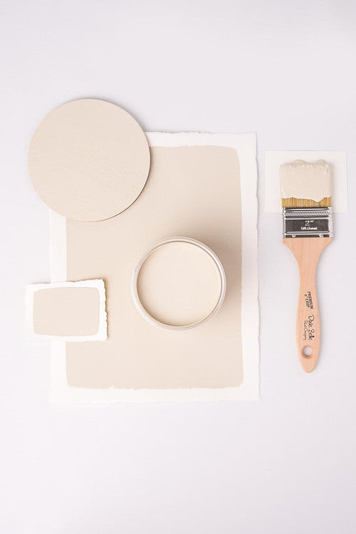 A flat lay image shows a beige paint theme with a round board, a paint can, and two swatches on white paper. A wooden-handled paintbrush with beige paint is placed to the right, contrasting the minimal white background.