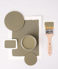 A jar of olive green paint is surrounded by painted swatches and wooden discs on a white background. A paintbrush with green paint is placed on the right side. The composition creates a balanced, artistic layout.