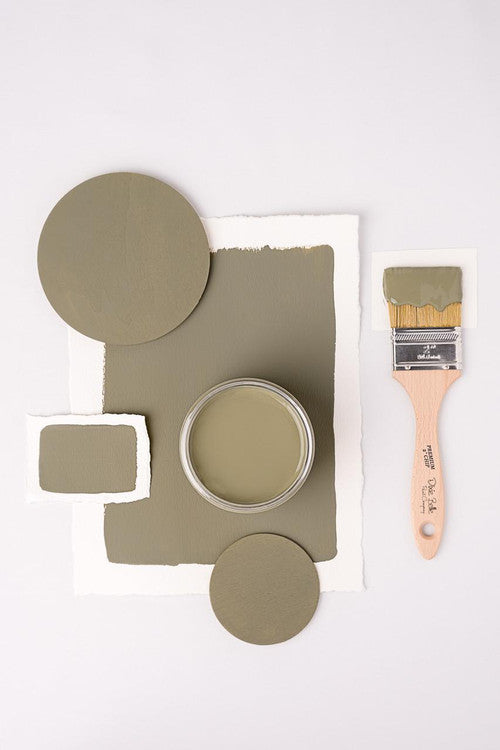 A jar of olive green paint is surrounded by painted swatches and wooden discs on a white background. A paintbrush with green paint is placed on the right side. The composition creates a balanced, artistic layout.