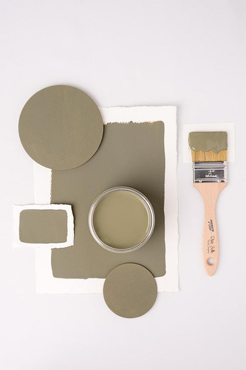 A jar of olive green paint is surrounded by painted swatches and wooden discs on a white background. A paintbrush with green paint is placed on the right side. The composition creates a balanced, artistic layout.