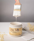 A hand holds a brush dripping with white paint above an open jar of Dixie Belle Chalk Mineral Paint in the color Gardenia. The jar is on a white surface with soft focus decorative elements around.