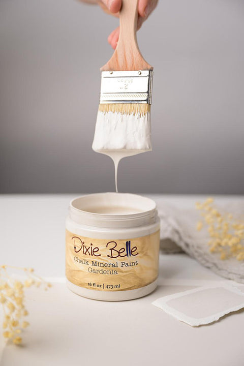A hand holds a brush dripping with white paint above an open jar of Dixie Belle Chalk Mineral Paint in the color Gardenia. The jar is on a white surface with soft focus decorative elements around.