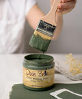 A person dips a paintbrush into an open jar of green Dixie Belle Chalk Mineral Paint labeled “Briar Patch.” The paintbrush is held above the jar, and green paint is spread on the surface below.