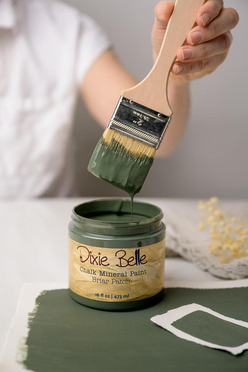 A person dips a paintbrush into an open jar of green Dixie Belle Chalk Mineral Paint labeled “Briar Patch.” The paintbrush is held above the jar, and green paint is spread on the surface below.