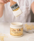 A person dips a paintbrush into a jar of Dixie Belle chalk mineral paint in Magnolia. The paint appears creamy and white, dripping from the brush. The jar is on a white surface with decorative elements nearby.