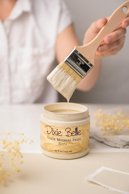 A person holds a brush coated with pale cream-colored paint above an open jar labeled "Dixie Belle Chalk Mineral Paint Ecru." The jar is on a white surface with dried flowers nearby. The background is softly blurred, focusing on the paint and brush.
