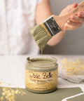 A person holds a paintbrush dripping with olive green paint above an open jar labeled "Dixie Belle Chalk Mineral Paint Thicket." The scene includes a green surface with small sprigs of yellow flowers and white paper in the background.