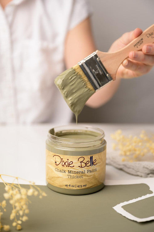 A person holds a paintbrush dripping with olive green paint above an open jar labeled "Dixie Belle Chalk Mineral Paint Thicket." The scene includes a green surface with small sprigs of yellow flowers and white paper in the background.