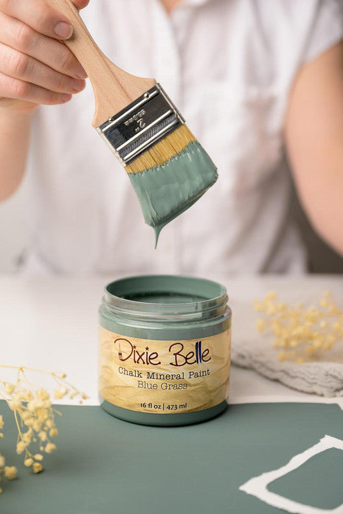 A person holds a paintbrush dripping with green paint above an open jar labeled "Dixie Belle Chalk Mineral Paint Blue Grass." The paintbrush is partially covered in matching green paint. Dry flowers and a fabric piece are nearby.