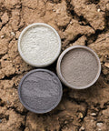 Three round containers filled with different shades of clay powder—light gray, medium gray, and dark gray—are arranged in a triangle on cracked, dry brown earth.
