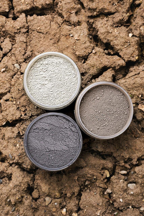 Three round containers filled with different shades of clay powder—light gray, medium gray, and dark gray—are arranged in a triangle on cracked, dry brown earth.