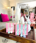 A wooden tray on a table holds pastel-colored Easter decorations, including a sign reading "Happy Easter," a ceramic bunny, candles, and colorful speckled eggs. A  dining room with pink chairs and decorative elements is in the background.