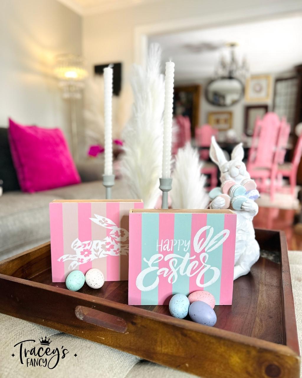 A wooden tray on a table holds pastel-colored Easter decorations, including a sign reading "Happy Easter," a ceramic bunny, candles, and colorful speckled eggs. A  dining room with pink chairs and decorative elements is in the background.