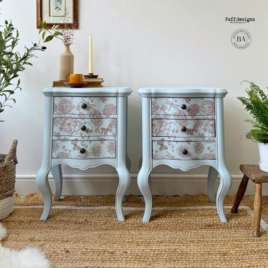 A pair of light blue vintage nightstands with three drawers each, adorned with floral patterns in light brownish red. The stands are placed on a woven rug, with potted plants, a candle, and books on top. A framed picture hangs on the wall above the left stand.
