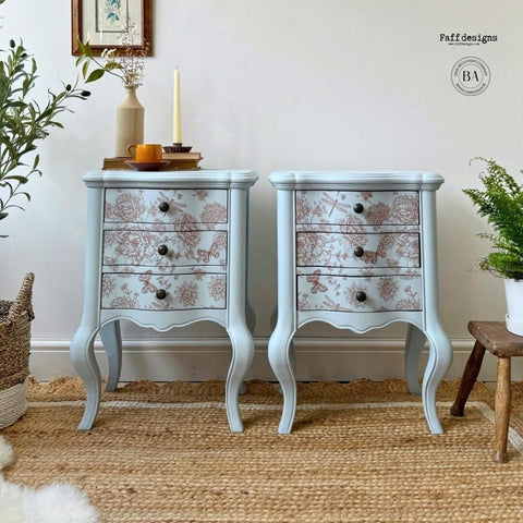 A pair of light blue vintage nightstands with three drawers each, adorned with floral patterns in light brownish red. The stands are placed on a woven rug, with potted plants, a candle, and books on top. A framed picture hangs on the wall above the left stand.