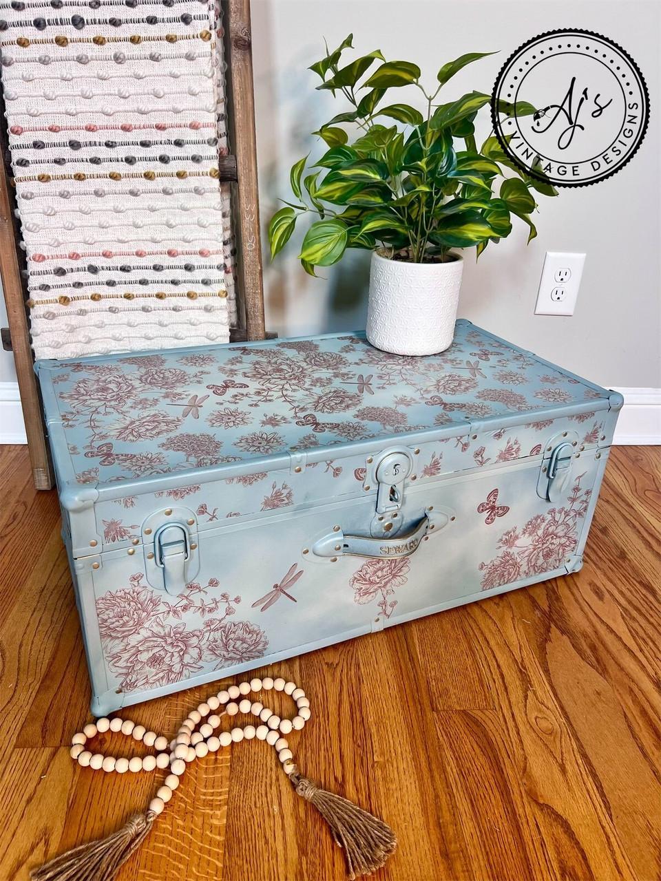 A decorated blue trunk with floral and dragonfly patterns holds a potted plant. A long, beaded wooden garland is arranged in front on the wooden floor. A rustic striped towel hangs from a ladder in the background, and a watermark logo reads "AJ's Vintage Designs".