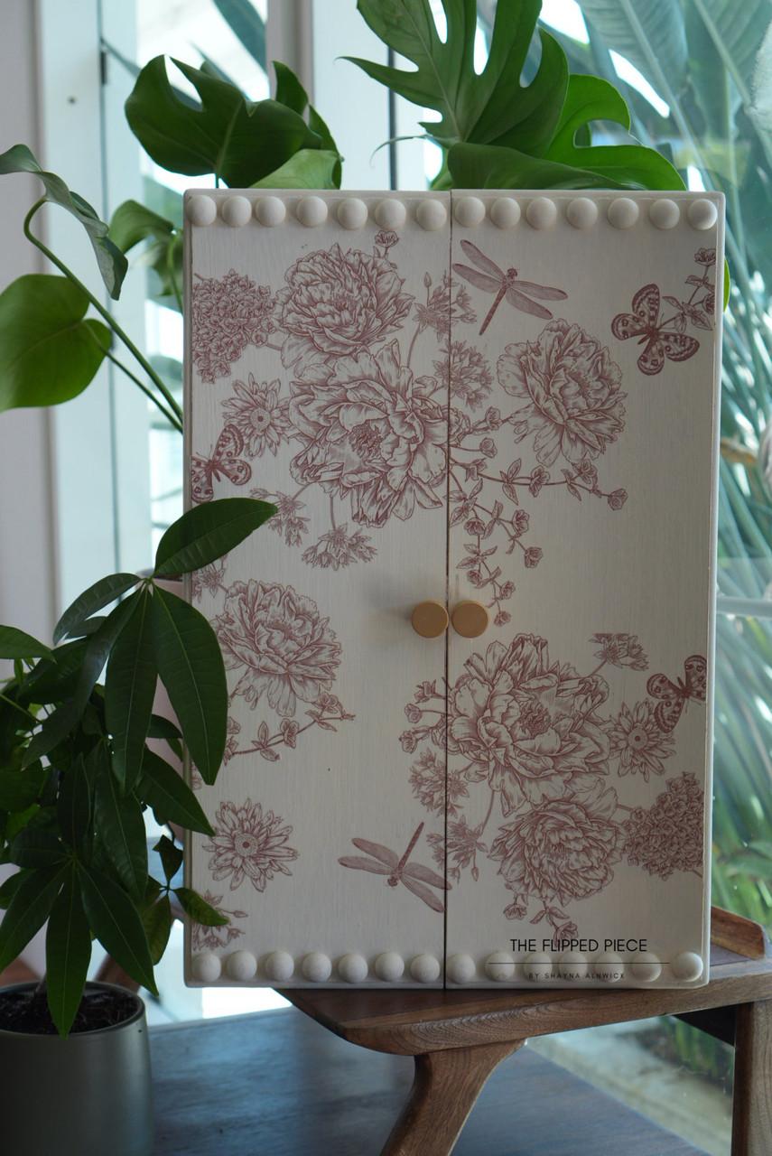 A decorative wooden cabinet with floral and insect patterns in red, featuring two beige knobs and a border of round wooden peg embellishments. It sits on a wooden stand, surrounded by lush green plants. Sunlight streams through a nearby window.