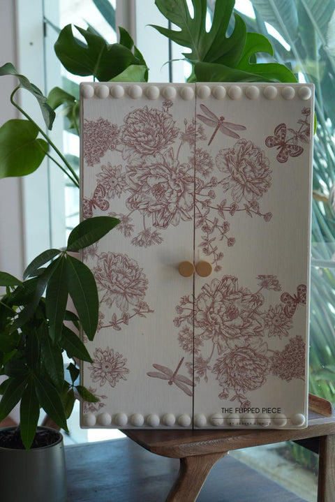 A decorative wooden cabinet with floral and insect patterns in red, featuring two beige knobs and a border of round wooden peg embellishments. It sits on a wooden stand, surrounded by lush green plants. Sunlight streams through a nearby window.