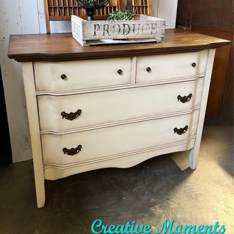 Dixie Belle Paint Drop Cloth Chalk Mineral Paint is the perfect paint for any DIY project! A vintage-style dresser with a dark wooden top and distressed cream-colored body featuring two small upper drawers and two large lower drawers. A wooden crate marked "Produce" and a small plant sit on top. Text reads "Creative Moments.