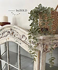A vintage-style wooden cabinet with ornate carvings, featuring a trailing potted plant and a candle on top. A wicker basket holds the plant, and the cabinet has glass-paneled doors. The setting appears rustic and elegant.
