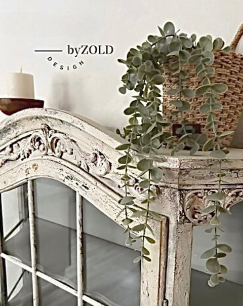 A vintage-style wooden cabinet with ornate carvings, featuring a trailing potted plant and a candle on top. A wicker basket holds the plant, and the cabinet has glass-paneled doors. The setting appears rustic and elegant.