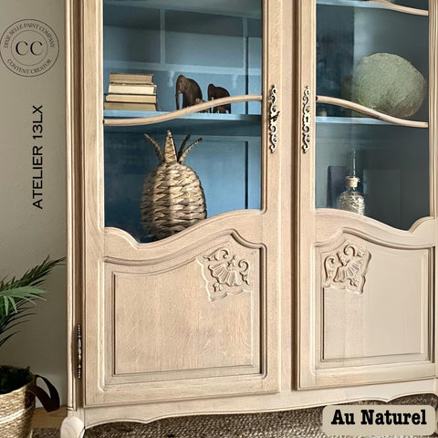 A rustic wooden cabinet with glass doors showcases decorative items, including stacked books, a woven basket, and small ornaments. The cabinet features ornate designs and is placed against a blue-painted interior background. Text reads "Au Naturel.