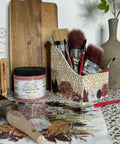 A decorative scene features art supplies on a surface. There are various paintbrushes in a floral-patterned holder, a jar of pink paint labeled 'Silk Desert Rose,' a flat wooden board, a ceramic vase, and a floral-patterned mat with scattered brushes and a small plant.