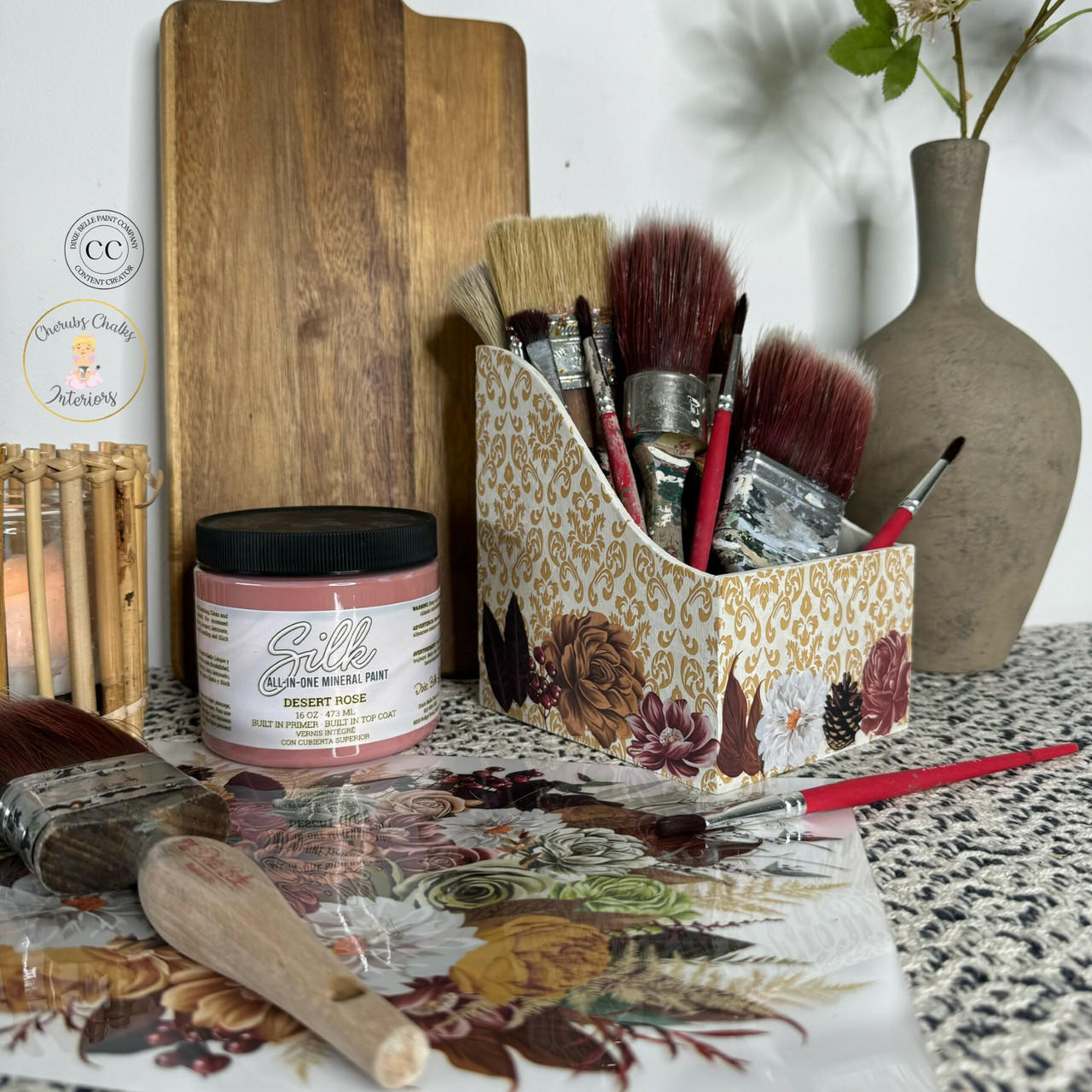 A decorative scene features art supplies on a surface. There are various paintbrushes in a floral-patterned holder, a jar of pink paint labeled 'Silk Desert Rose,' a flat wooden board, a ceramic vase, and a floral-patterned mat with scattered brushes and a small plant.