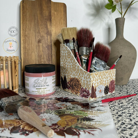 A decorative scene features art supplies on a surface. There are various paintbrushes in a floral-patterned holder, a jar of pink paint labeled 'Silk Desert Rose,' a flat wooden board, a ceramic vase, and a floral-patterned mat with scattered brushes and a small plant.