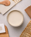 A top-down view of an open beige-colored jar on a woven textured surface. Nearby are a wooden spoon, a square ceramic tile, a woven mat, a piece of paper, and a woven straw item. Neutral and earthy tones predominate the scene.