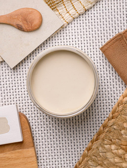 A top-down view of an open beige-colored jar on a woven textured surface. Nearby are a wooden spoon, a square ceramic tile, a woven mat, a piece of paper, and a woven straw item. Neutral and earthy tones predominate the scene.