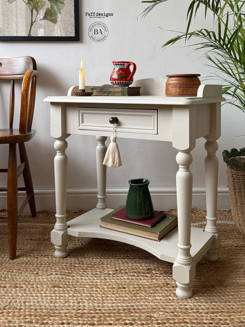 A vintage-style cream side table with a decorative tassel handle, holding a lit candle, red mug, and small brown container. Below, a green pitcher rests on stacked books. The setup is on a textured rug with a chair and potted plant nearby.