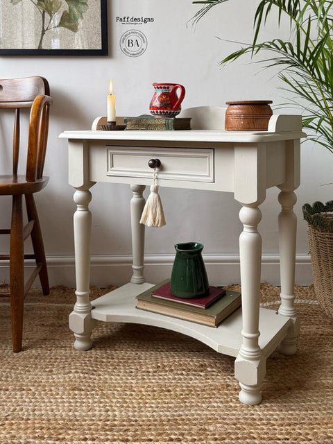 A vintage-style cream side table with a decorative tassel handle, holding a lit candle, red mug, and small brown container. Below, a green pitcher rests on stacked books. The setup is on a textured rug with a chair and potted plant nearby.