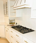 A modern white kitchen with gold hardware, a gas stovetop, white cabinets, open shelving, a vent hood with gold trim, a small plant, and neatly arranged condiments on the counter. The MTM logo is visible in the corner.