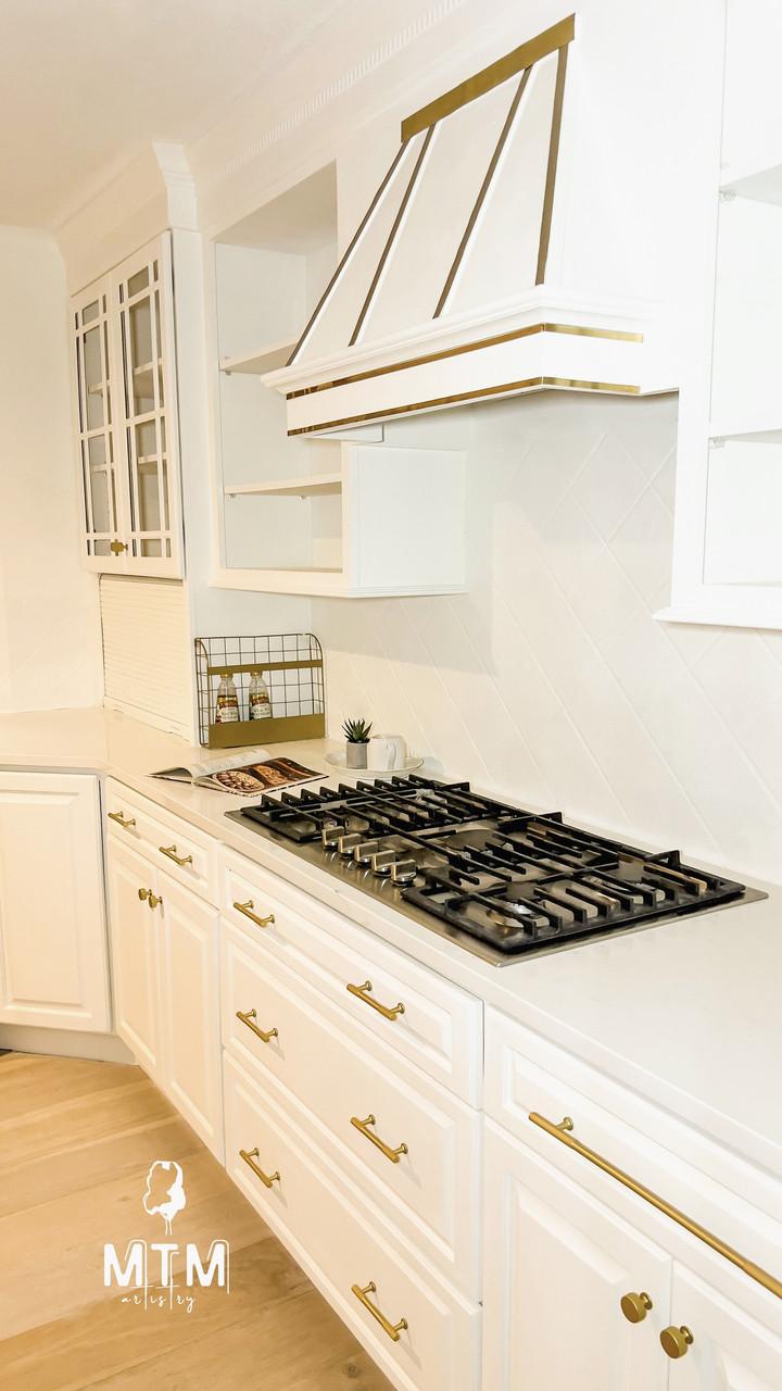 A modern white kitchen with gold hardware, a gas stovetop, white cabinets, open shelving, a vent hood with gold trim, a small plant, and neatly arranged condiments on the counter. The MTM logo is visible in the corner.