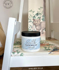 A white chair with a floral seat cushion holds a jar of Dixie Belle Silk all-in-one mineral paint. Behind the jar, thereâ€™s an image of a bear and trees. A round â€œcontent creatorâ€ badge is visible in the top left corner.
