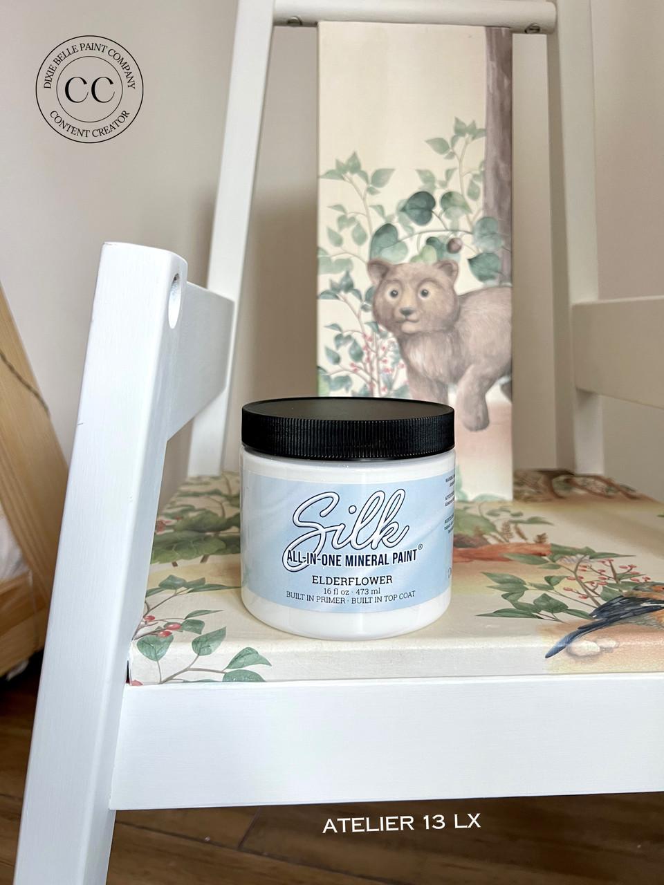 A white chair with a floral seat cushion holds a jar of Dixie Belle Silk all-in-one mineral paint. Behind the jar, thereâ€™s an image of a bear and trees. A round â€œcontent creatorâ€ badge is visible in the top left corner.