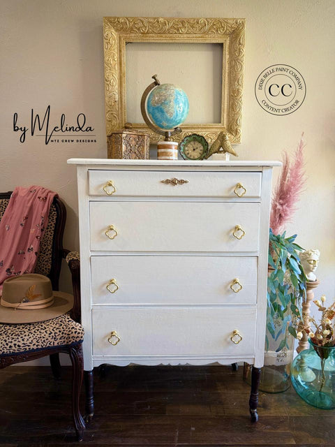 A white vintage dresser with gold handles stands against a wall, topped with a globe, books, a small clock, and a decorative frame. A leopard-print chair with a hat and a pink scarf and a vase with pampas grass are nearby.