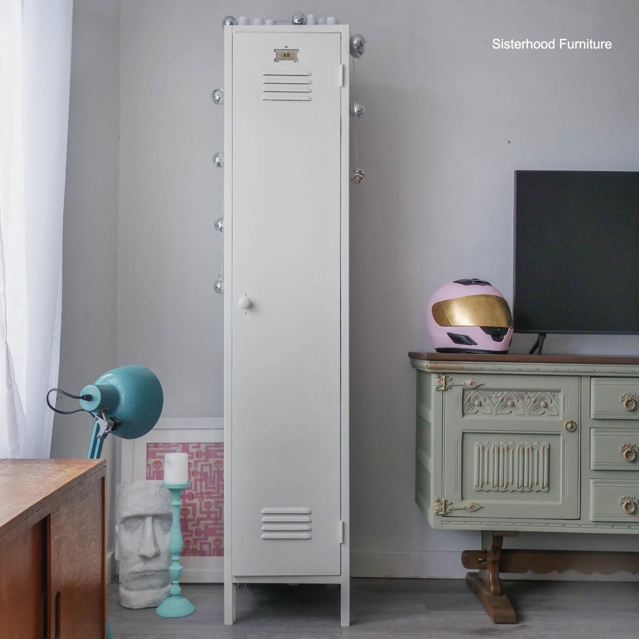 A tall, white metal locker stands between a blue lamp, a sculpted head, and a mint green vintage dresser. A pink helmet with a gold visor sits on the dresser beside a TV. The room has a modern, eclectic style.