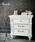 A white vintage nightstand with gold handles stands against a dark, ornate wallpaper. On top are books, a purse, and toiletries. A decorative black wire mannequin stands to the left. "Theresa Lee NTS Design Co." watermark is in the corner.